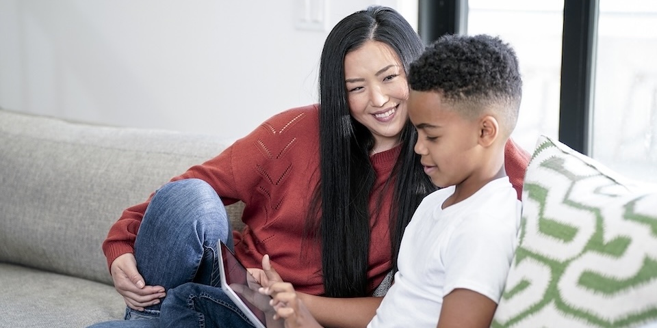 Mom and son sitting on couch. Mom smiles and son works on tablet.